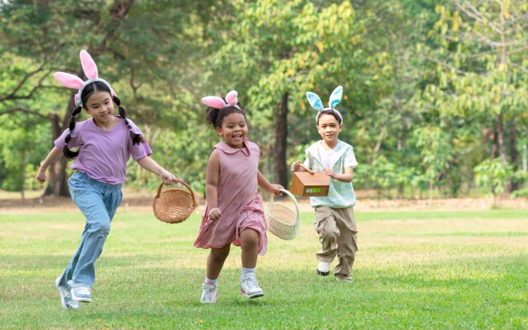 Three children wearing bunny ears run across a grassy park during an Easter egg hunt, carrying baskets and a small wooden box.