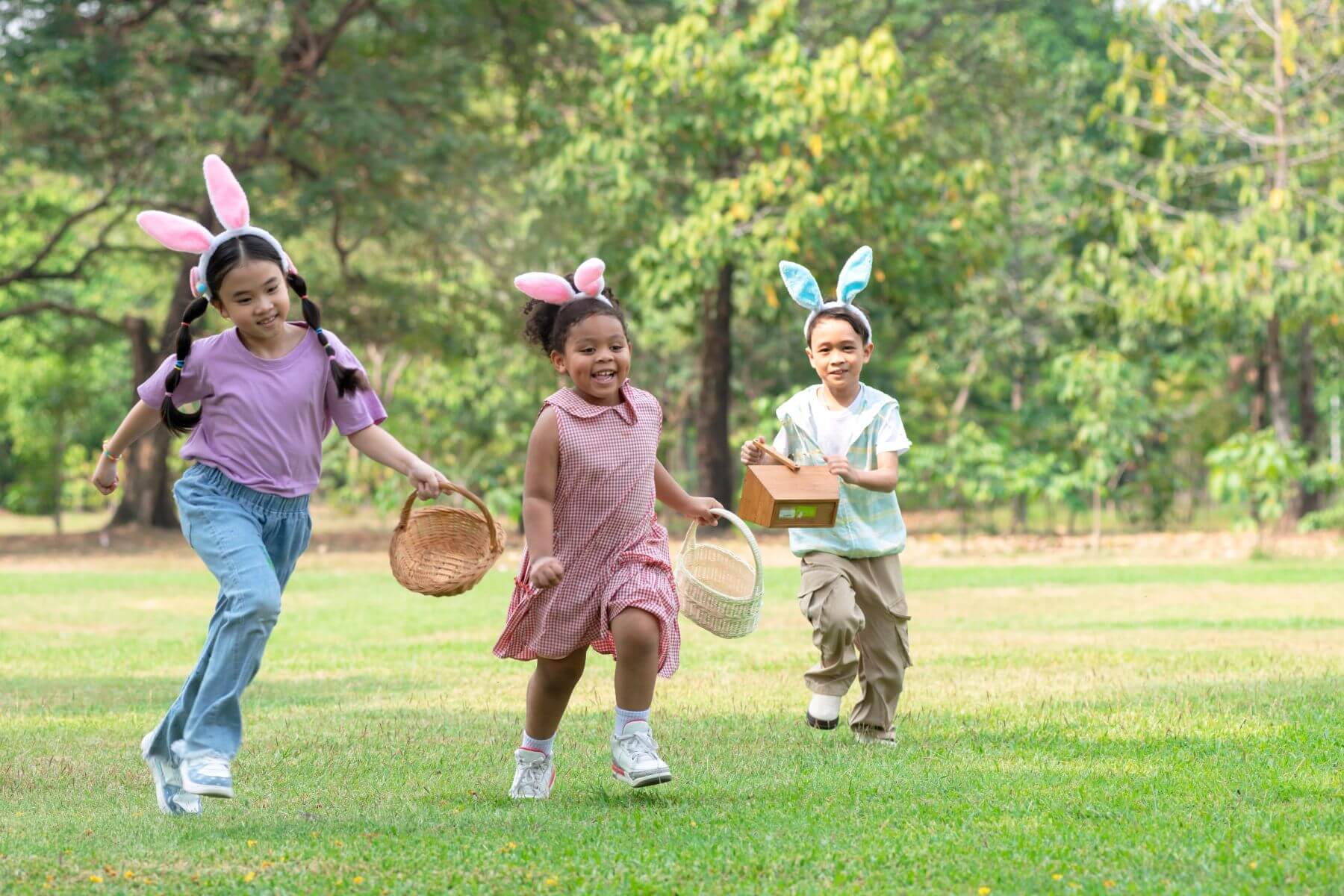 Three children wearing bunny ears run across a grassy park during an Easter egg hunt, carrying baskets and a small wooden box.