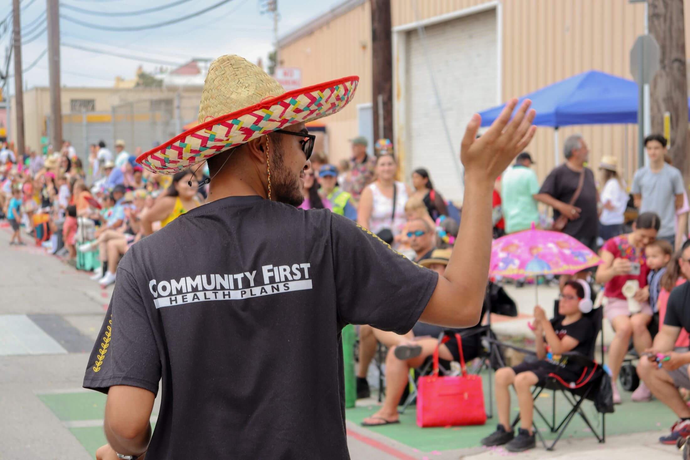 Man in a sombrero and "Community First Health Plans" shirt waves to spectators lining a street during an outdoor festival or parade.