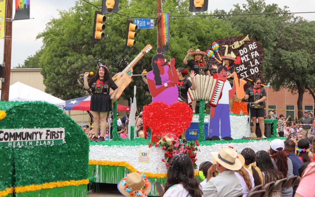 A parade float decorated with green foliage for Community First Health Plans features performers celebrating Mexican culture with large cardboard cutouts of musical instruments and figures in front of cheering spectators.