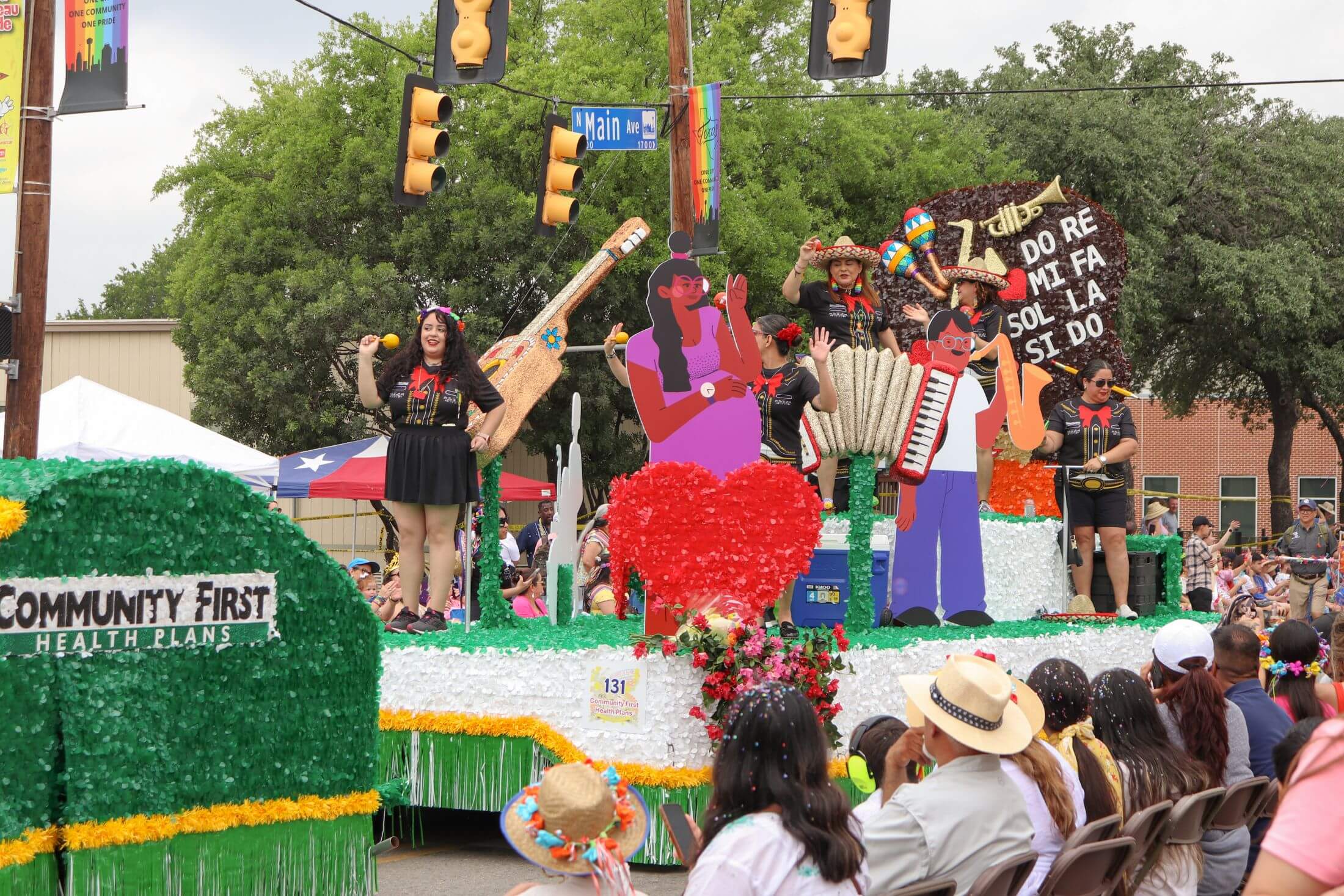A parade float decorated with green foliage for Community First Health Plans features performers celebrating Mexican culture with large cardboard cutouts of musical instruments and figures in front of cheering spectators.