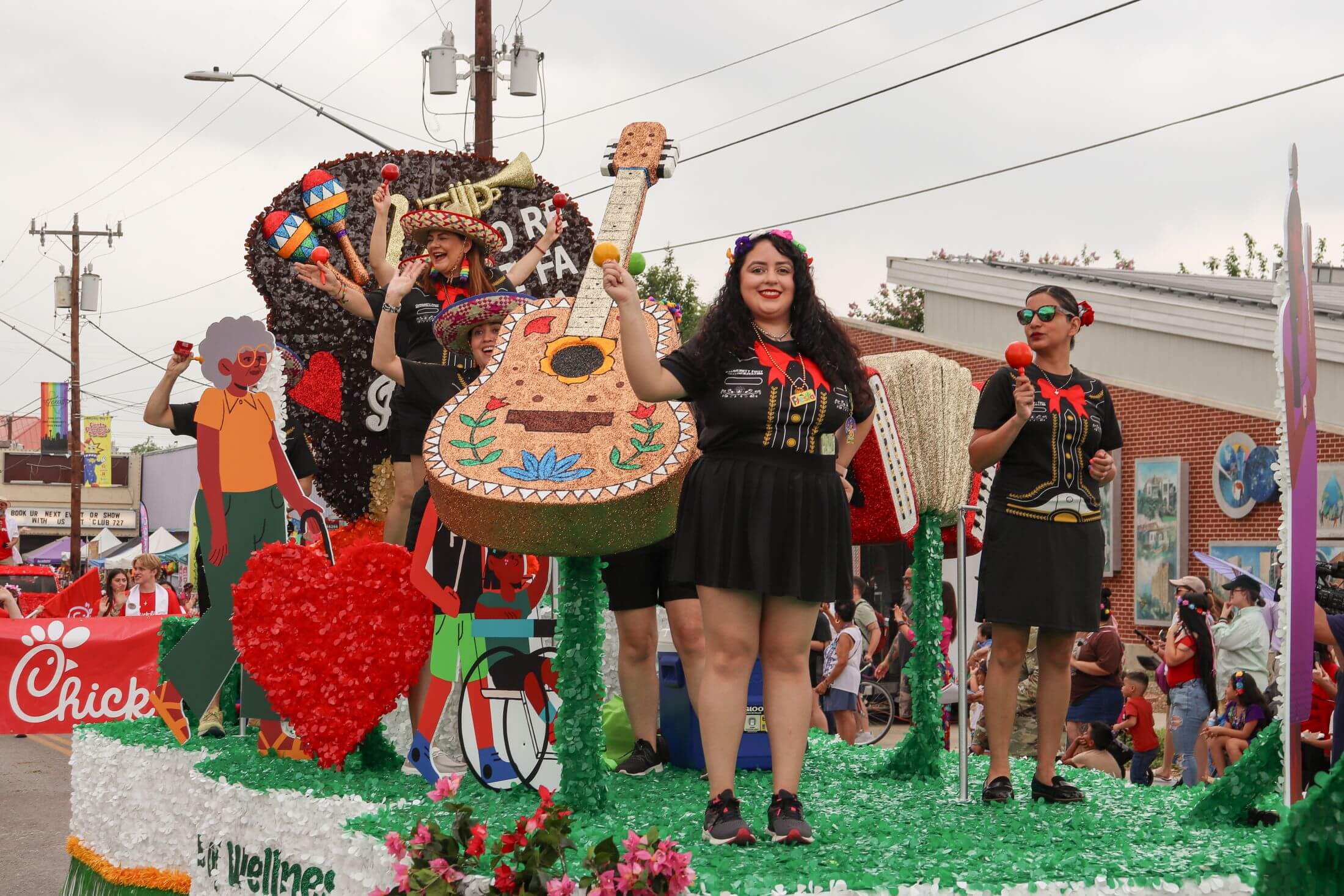 Women wearing festive attire participate in the Battle of the Flowers parade on a float decorated with a large guitar, maracas, and heart shapes.