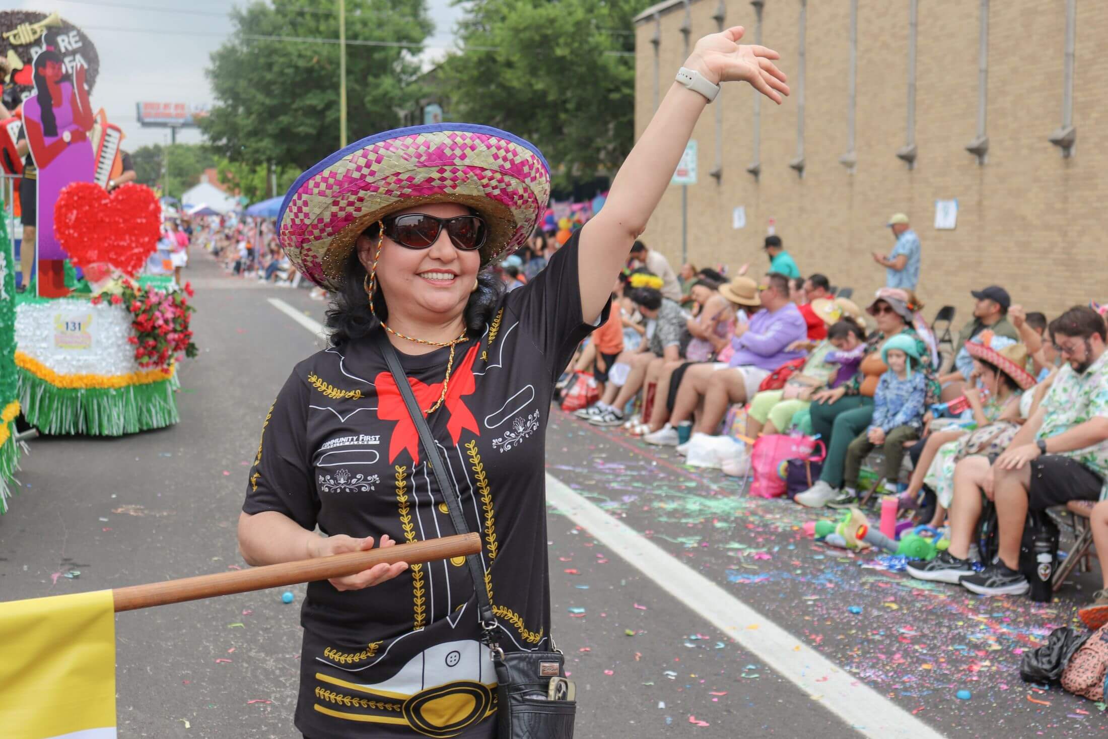 Woman in a sombrero and mariachi-style shirt waves from a parade, holding a flag pole while spectators sit along the street littered with confetti.