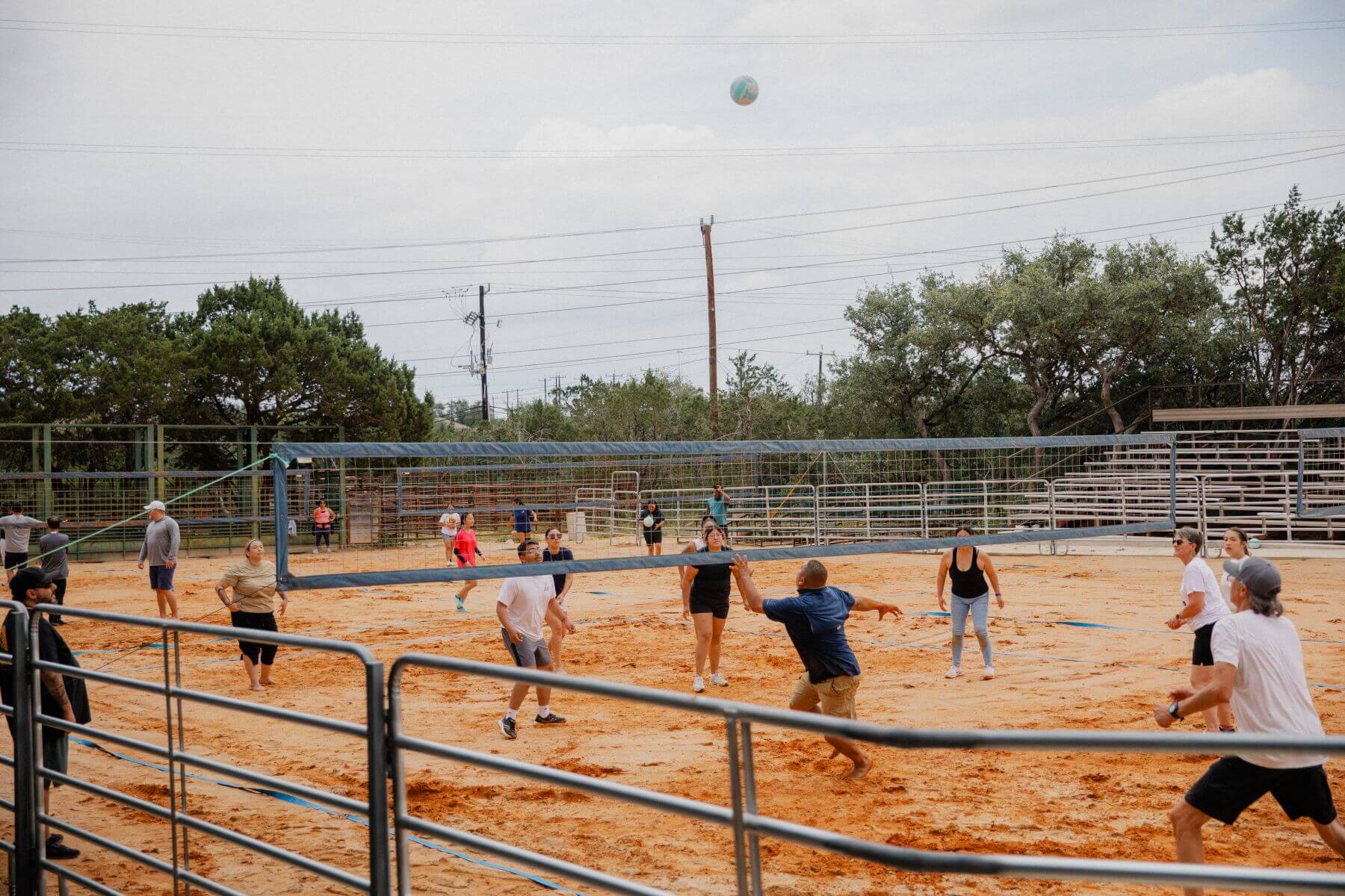 A group of adults plays volleyball on a sandy court enclosed by metal fencing under an overcast sky with power lines visible above the surrounding trees.