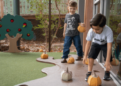 Two young boys play outdoors with small decorative pumpkins on a textured brown path beside green turf and a wooden apple tree cutout.