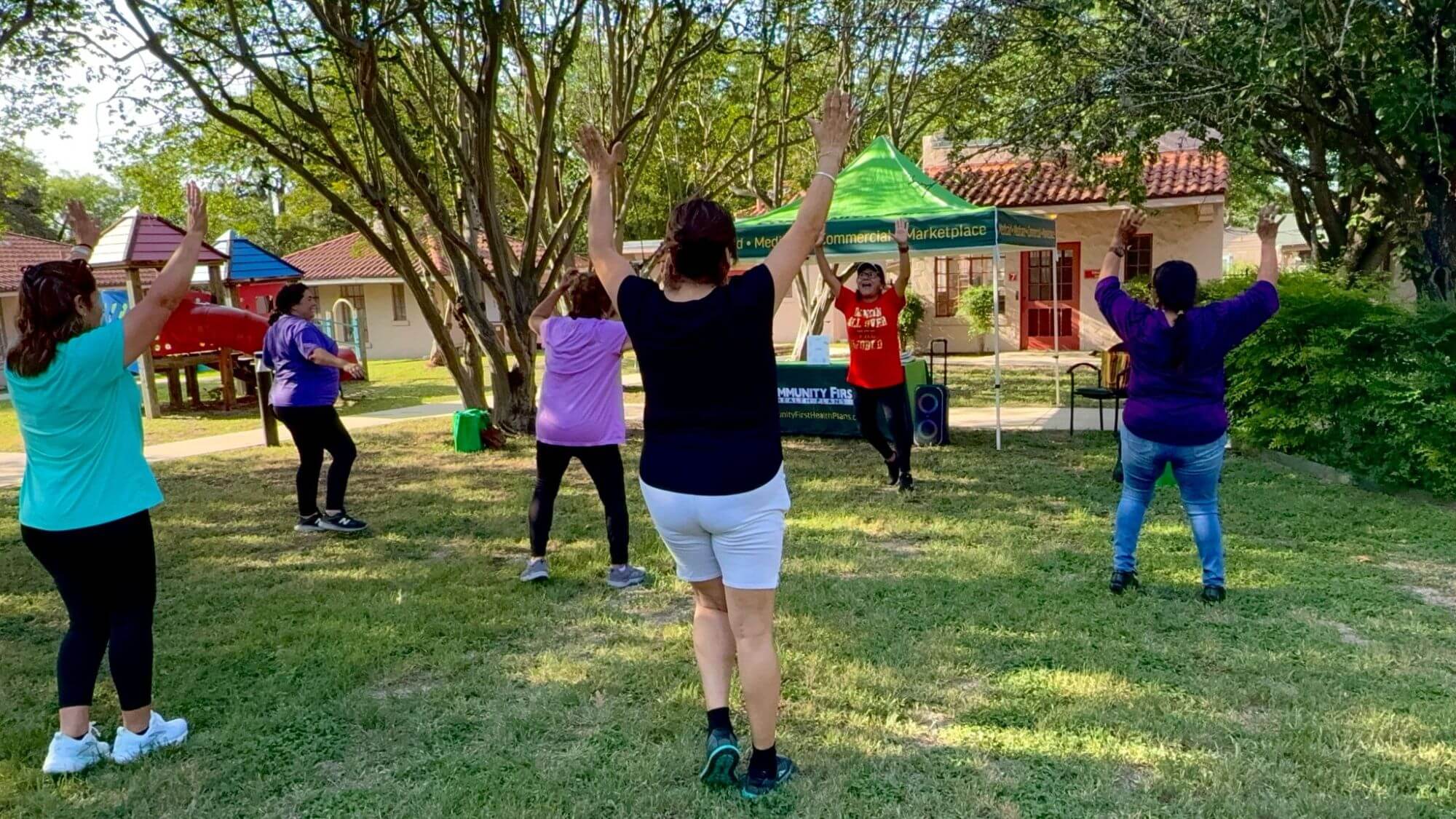A group of diverse adults exercises outdoors on a sunny day with their arms raised, led by an instructor near a green canopy tent.