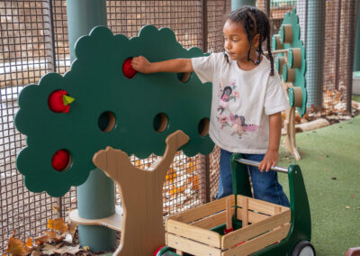 Young girl interacts with a green wooden apple-picking play tree structure next to a toy wagon on artificial turf.