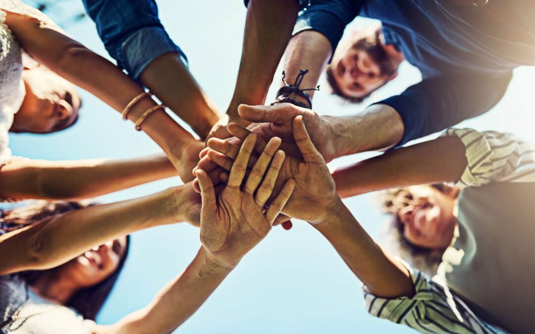Low-angle shot of a diverse group of people stacking hands together in a circle outdoors against a bright blue sky.