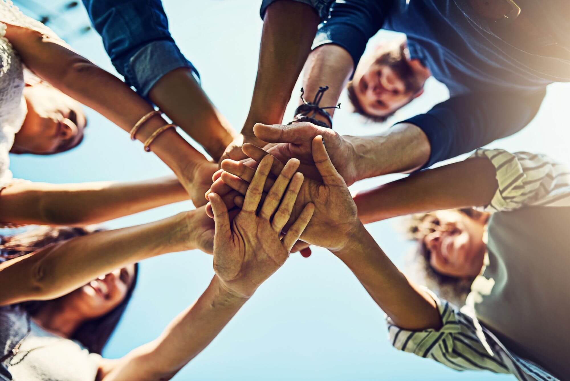 Low-angle shot of a diverse group of people stacking hands together in a circle outdoors against a bright blue sky.
