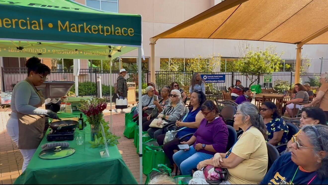 A woman demonstrates cooking outdoors under a green tent labeled "Marketplace" while an audience, mostly older women, watches seated nearby.