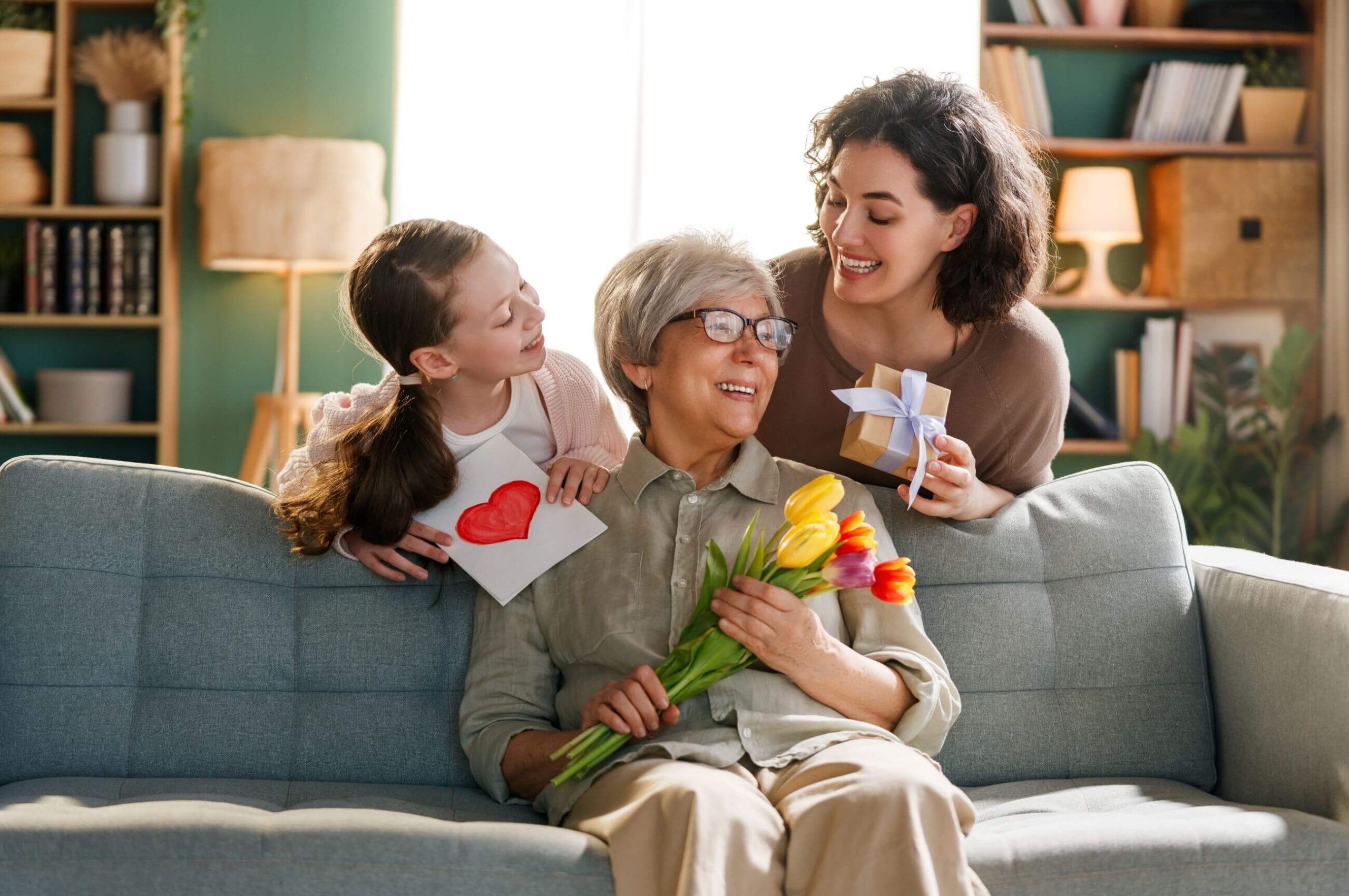 Grandmother smiling as she receives colorful tulips, a handmade card, and a gift from her daughter and granddaughter in a cozy living room, capturing a warm, multigenerational Mother’s Day moment.