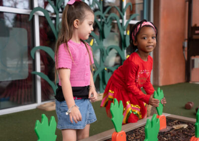 Two young girls play in a garden area with oversized toy carrots, one standing in a pink cardigan and denim skirt, the other kneeling in a red dress.