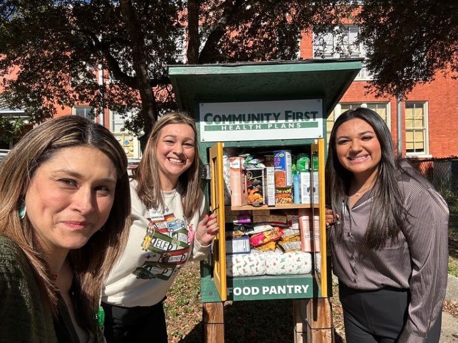 Three smiling women stand outdoors next to a small, stocked community first health plans food pantry with a red brick building in the background.