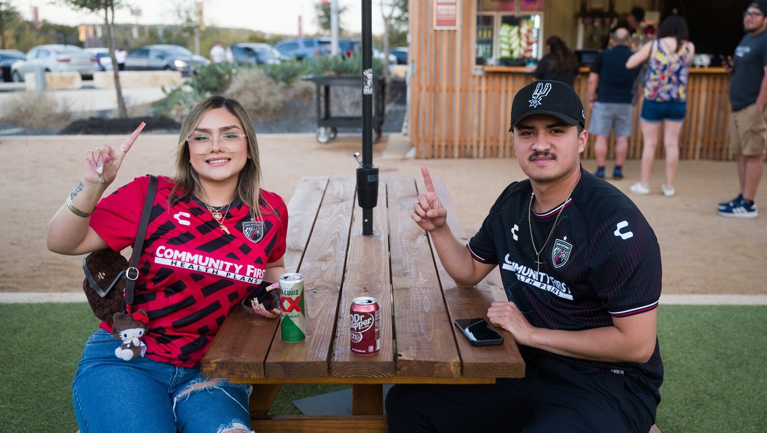 A young couple wearing soccer jerseys sits at an outdoor wooden picnic table with canned drinks while flashing a number one sign.