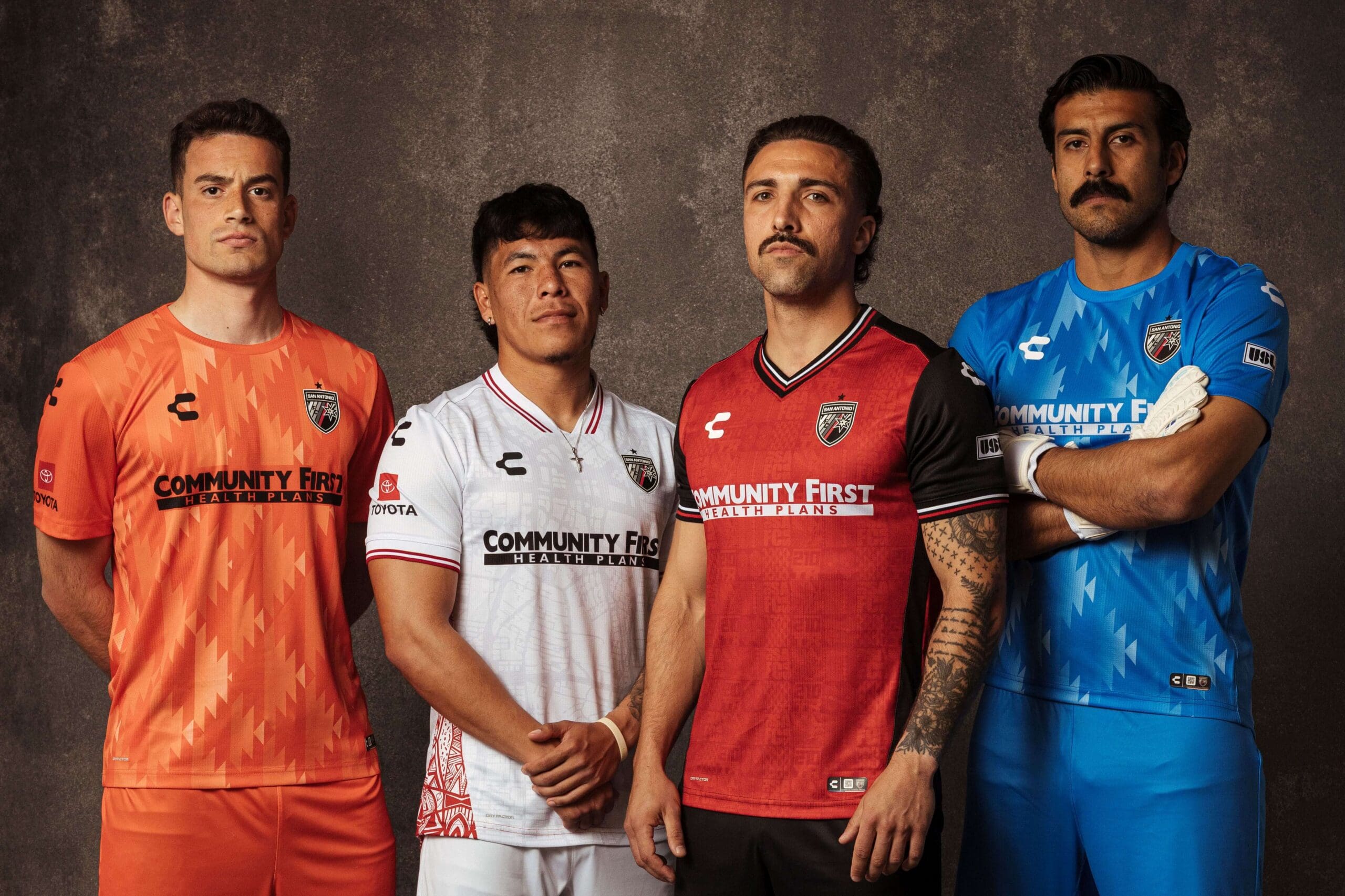 Four male soccer players stand against a dark background modeling orange, white, red, and blue San Antonio FC jerseys sponsored by Community First Health Plans.