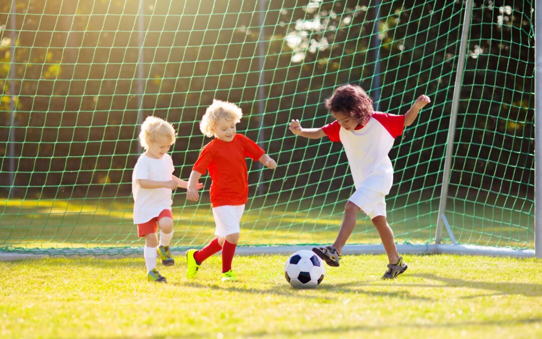 Children playing soccer in a grassy park, kicking a ball in front of a goal net during a sunny day.