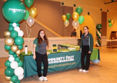 Two women stand at a table with "COMMUNITY FIRST HEALTH PLANS" branding, surrounded by green, gold, and white balloons.