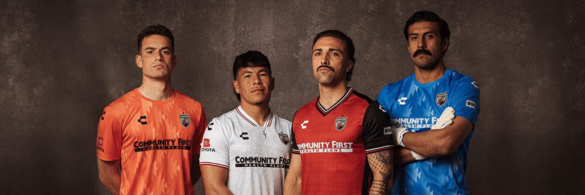 Four male soccer players stand against a dark background modeling orange, white, red, and blue San Antonio FC jerseys sponsored by Community First Health Plans.