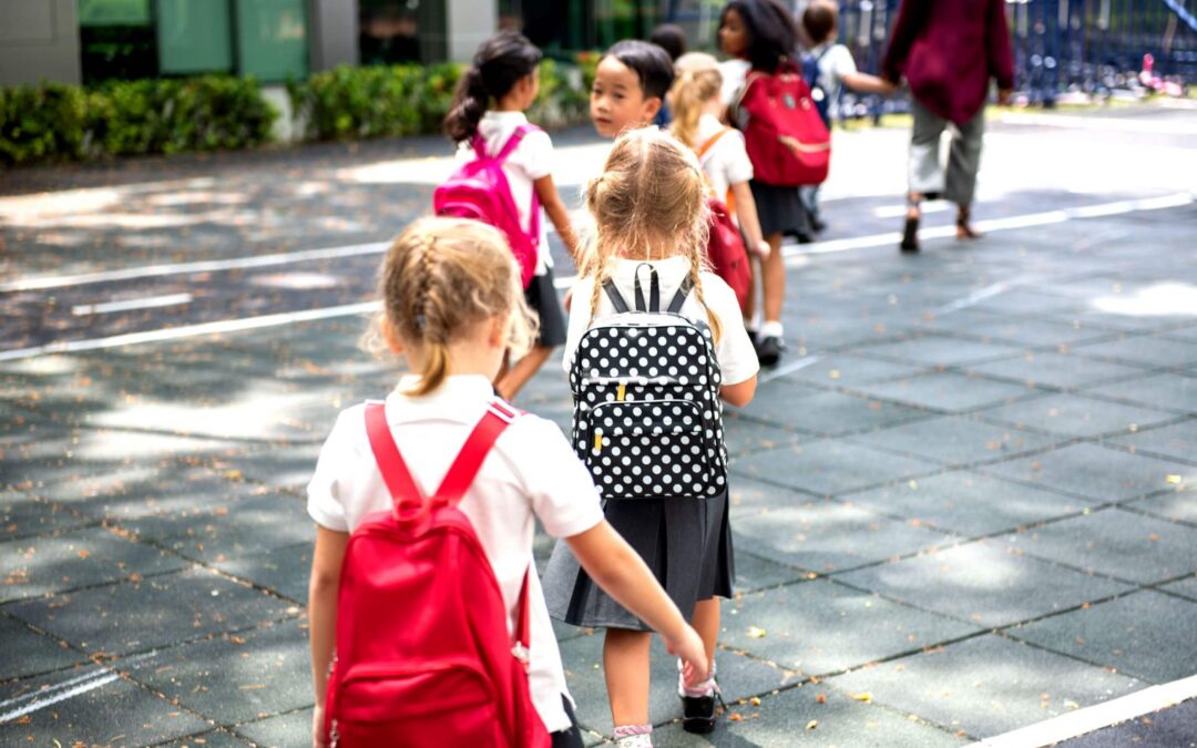 Children with red backpacks walk in a line on a playground. They wear school uniforms and appear to be in an outdoor school setting on a sunny day.