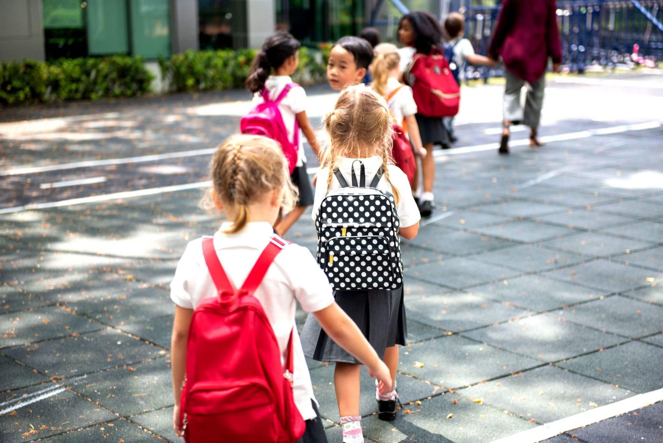 Children with red backpacks walk in a line on a playground. They wear school uniforms and appear to be in an outdoor school setting on a sunny day.