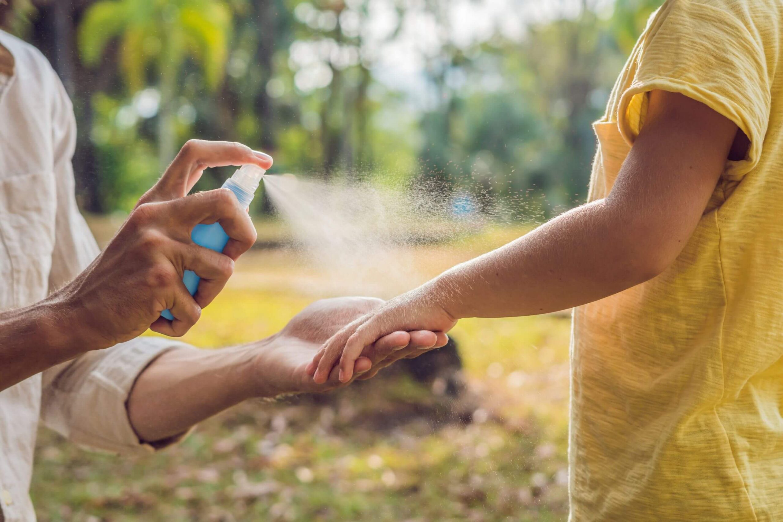 A person sprays insect repellent onto another person's arm outdoors.