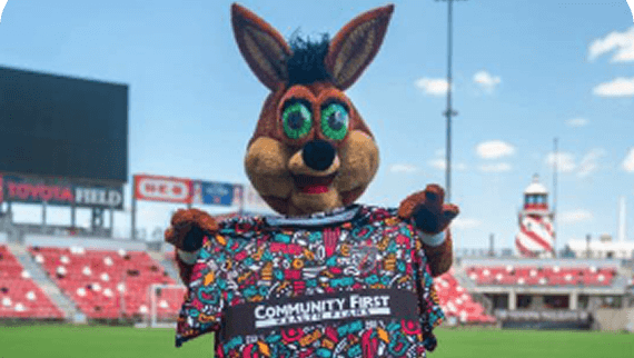The Coyote stands on a soccer field holding up a colorful shirt with "Community First" written on it.